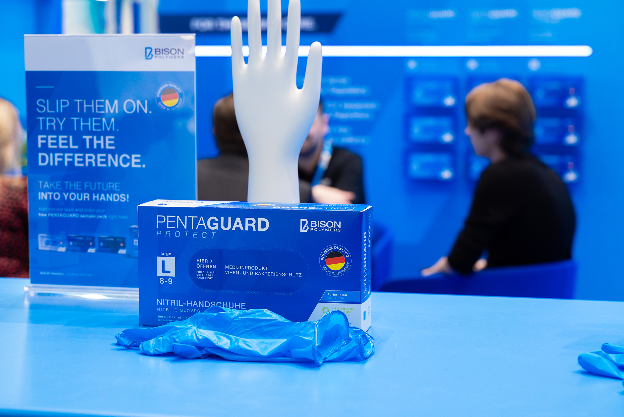 PENTAGUARD Nitril-Einmalhandschuhe von BISON Polymers auf der MEDICA: Im Vordergrund eine Produktbox, ein Paar Handschuhe sowie eine Keramikhand zur Passform-Demonstration; im Hintergrund ein Messegespräch.