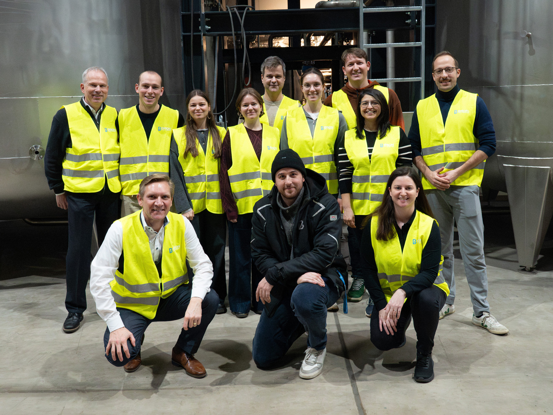 Group photo of medical students and BISON Polymers staff wearing yellow safety vests in a production hall in front of two large metal tanks.