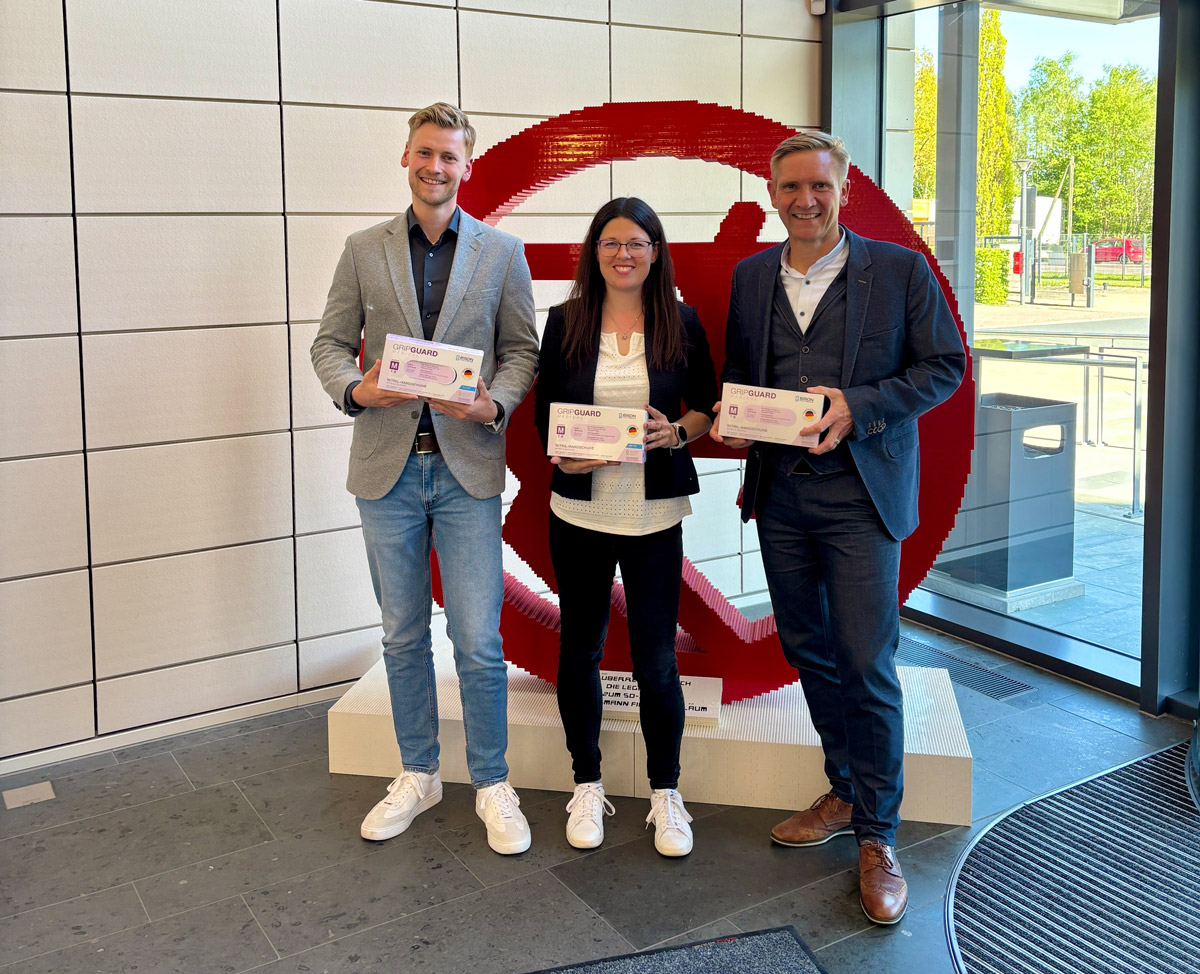 Sascha Schellenberg, Carina Kirchhoff und Jan Schirle präsentieren Handschuhboxen von BISON Polymers vor einem großen Rossmann-Logo in der Rossmann-Zentrale in Burgwedel.